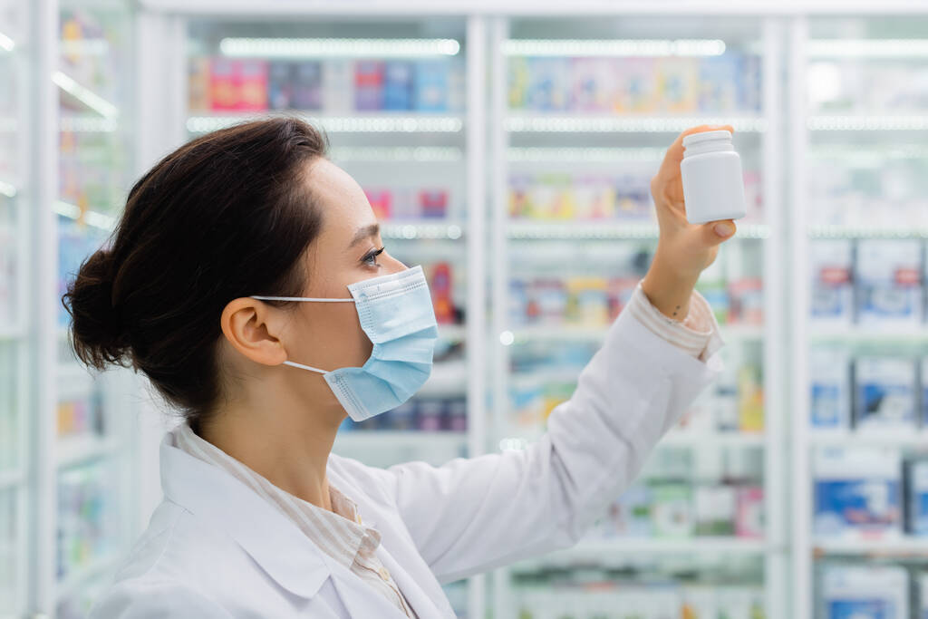 vista lateral del boticario morena en máscara médica mirando botella en farmacia - Foto, imagen