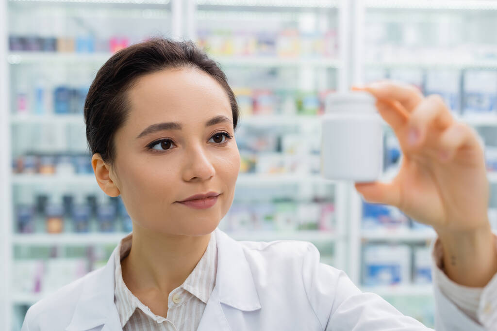 Аптекарь брюнетки в белом халате смотрит на размытую бутылку в аптеке - Фото, изображение