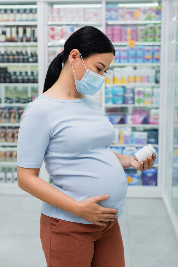 embarazada asiático cliente en médico máscara buscando en botella con vitaminas en droguería - Foto, imagen