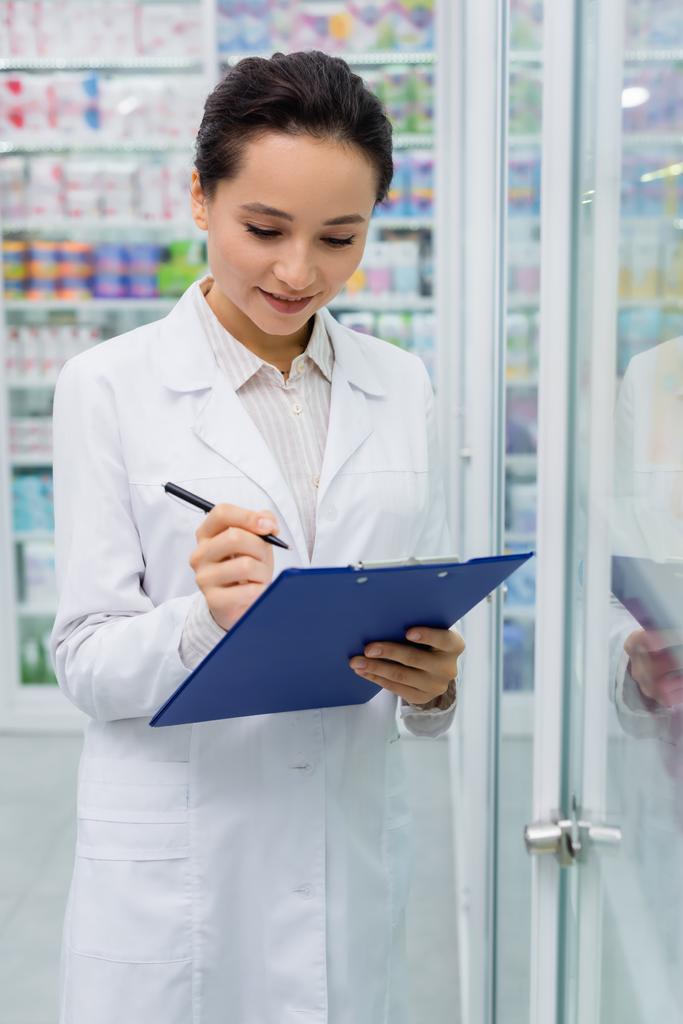 cheerful pharmacist in white coat writing on clipboard  - Photo, Image