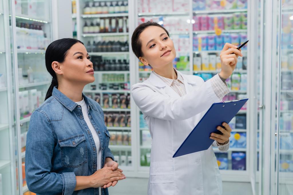 gyógyszerész tollal mutogat, miközben írótáblát tart ázsiai nő közelében a gyógyszertárban - Fotó, kép