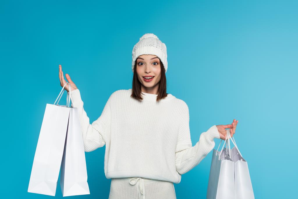 Mulher excitada em chapéu de malha e suéter segurando sacos de compras isolados em azul  - Foto, Imagem
