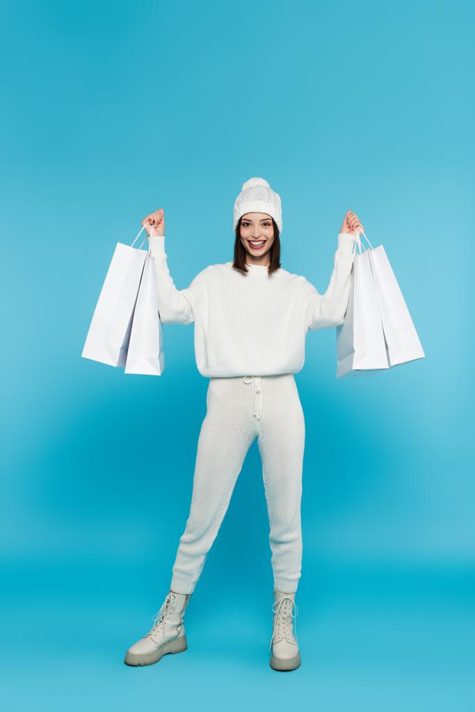 Cheerful shopaholic in sweater and hat holding shopping bags on blue background  - Photo, Image