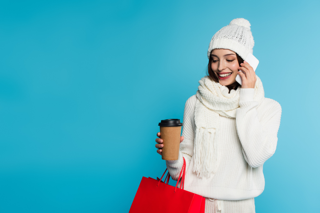 Sıcak giysiler içinde gülümseyen, akıllı telefondan konuşan, alışveriş torbaları tutan ve mavi üzerine izole edilmiş kahve içen kadın.  - Fotoğraf, Görsel