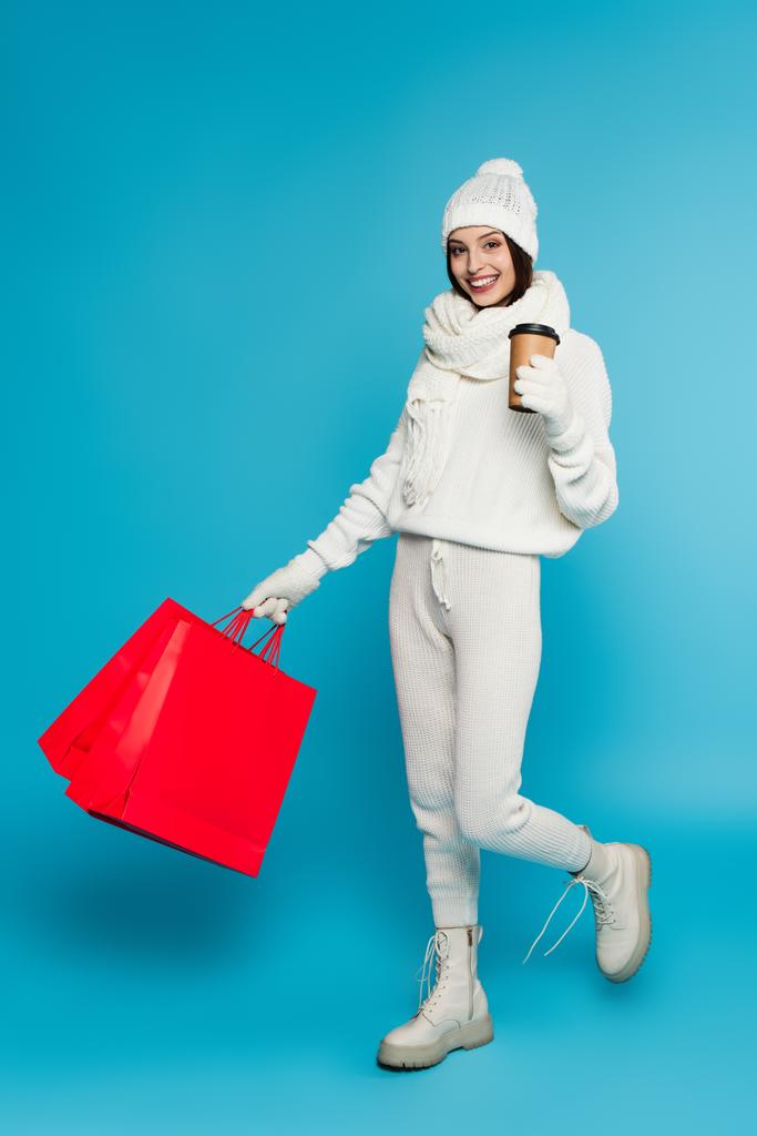 Χαρούμενη γυναίκα με πλεκτά ρούχα και γάντια που κρατά χάρτινο κύπελλο και τσάντες για ψώνια σε μπλε φόντο  - Φωτογραφία, εικόνα