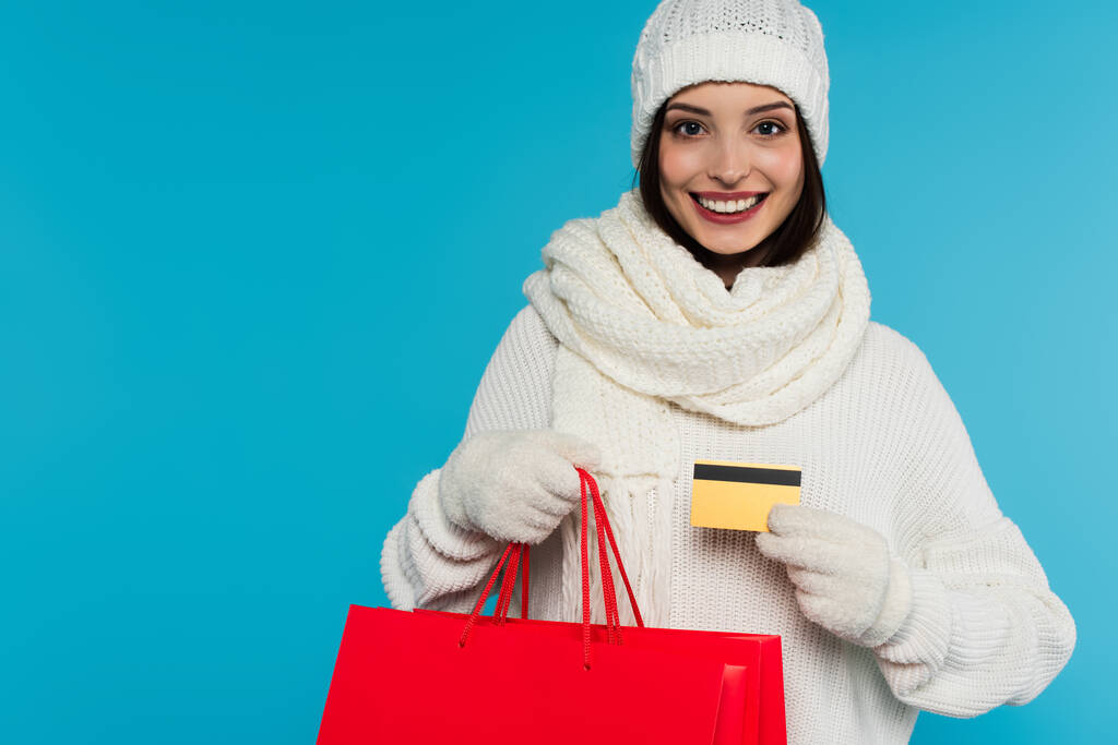 Jolie femme en chapeau et gants tenant des sacs à provisions et une carte de crédit isolée sur bleu  - Photo, image