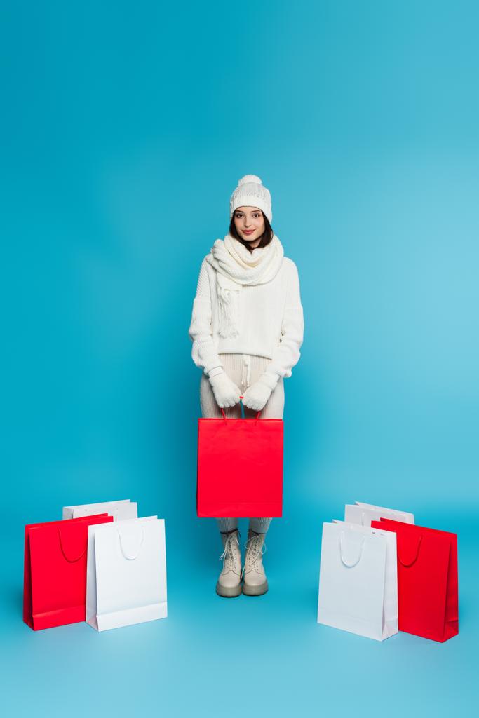 Pretty shopaholic in knitted clothes standing near shopping bags on blue background  - Photo, Image