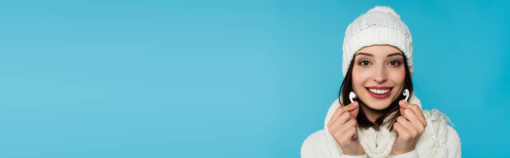 Junge Frau in warmer Kleidung lächelt, während sie kabellose Kopfhörer auf blauem Banner hält  - Foto, Bild