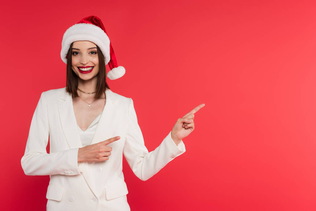 Femme gaie en chapeau de Père Noël et veste blanche pointant avec les doigts isolés sur rouge - Photo, image