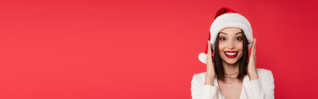 Захоплена жінка в капелюсі Санта дивиться на камеру ізольовано на червоному, банер
  - Фото, зображення