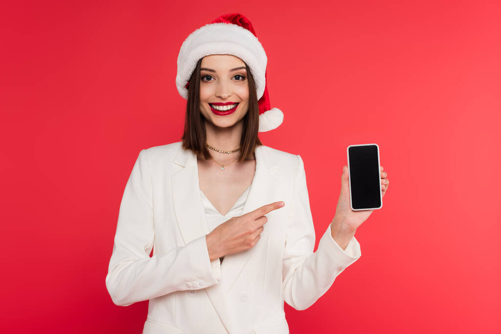 Noel Baba şapkalı neşeli kadın kırmızı ekranda boş ekranlı akıllı telefonu işaret ediyor.  - Fotoğraf, Görsel