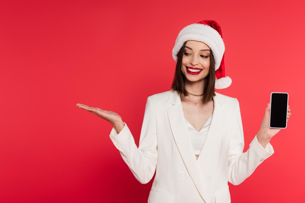 Noel Baba şapkalı şık bir kadın cep telefonunu boş ekran ile tutuyor ve eli kırmızı üzerine izole edilmiş bir şekilde gösteriyor.  - Fotoğraf, Görsel