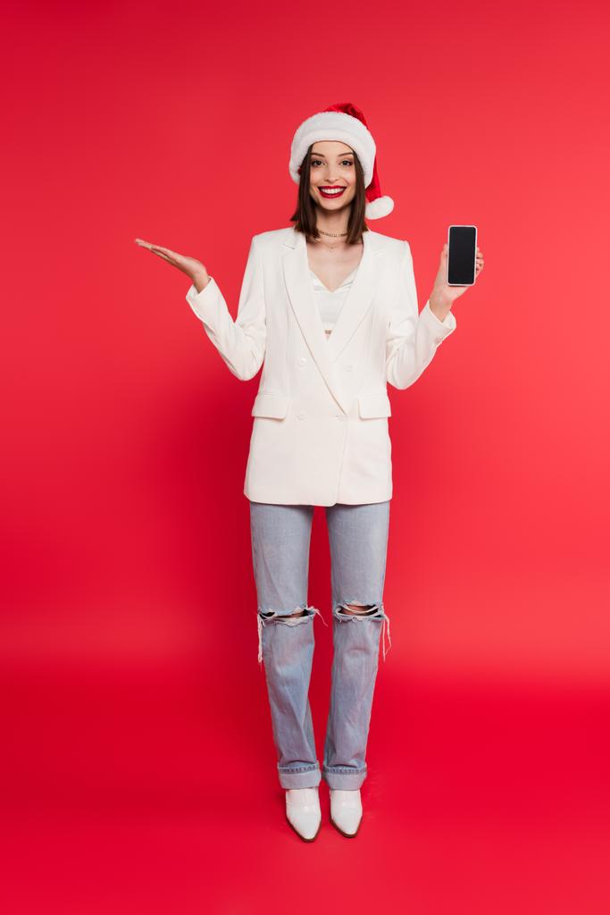 Fröhliche Frau mit Weihnachtsmannmütze und weißer Jacke hält Smartphone mit leerem Bildschirm und zeigt mit der Hand auf rotem Hintergrund  - Foto, Bild