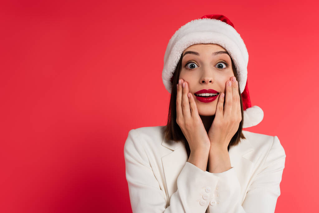 Astonished woman with red lips and santa hat looking at camera isolated on red  - Photo, Image