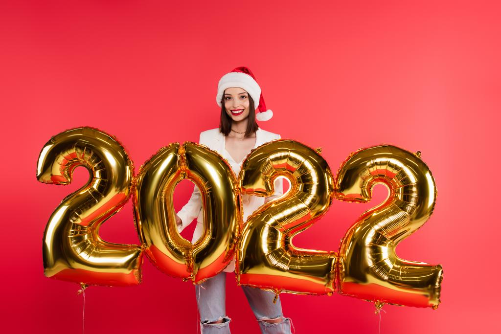 Ładna kobieta w kapeluszu Mikołaja stojąca w pobliżu balonów w kształcie liczb 2022 odizolowanych na czerwono  - Zdjęcie, obraz