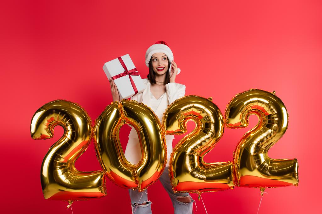 Uśmiechnięta kobieta w kapeluszu Mikołaja rozmawia na smartfonie i trzyma prezent w pobliżu balonów w kształcie 2022 numery izolowane na czerwono  - Zdjęcie, obraz