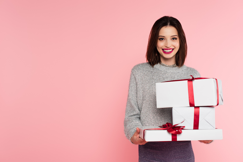 Hübsche Frau lächelt in die Kamera, während sie Geschenkboxen isoliert auf rosa hält  - Foto, Bild
