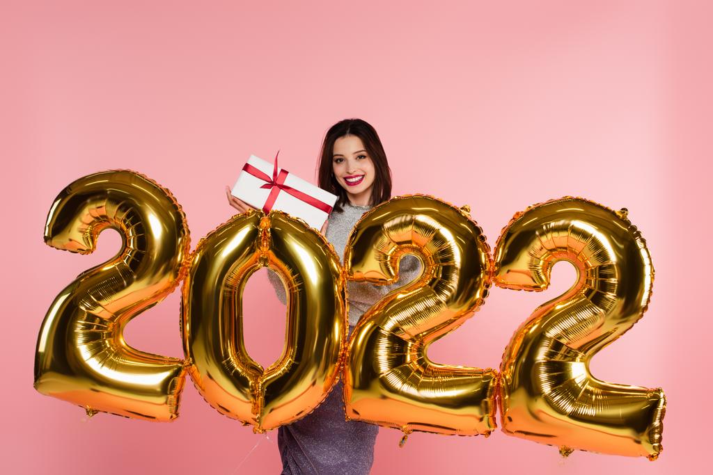 Hymyilevä nainen, jolla on lahjapakkaus lähellä ilmapalloja, 2022 numeroa, jotka on eristetty vaaleanpunaiselle  - Valokuva, kuva