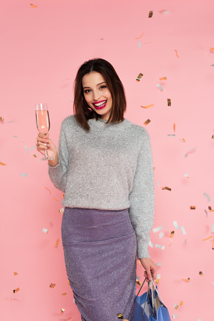 Trendy woman with champagne and shopping bags standing under confetti on pink background - Photo, Image