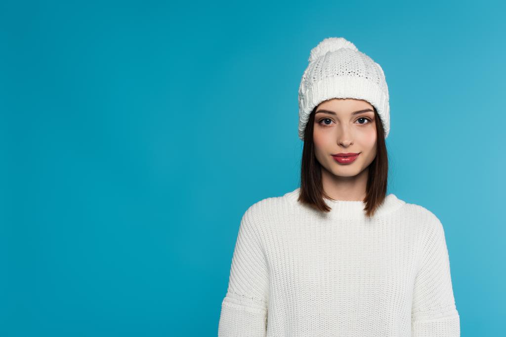Mujer bastante joven en sombrero blanco y suéter mirando a la cámara aislada en azul  - Foto, imagen