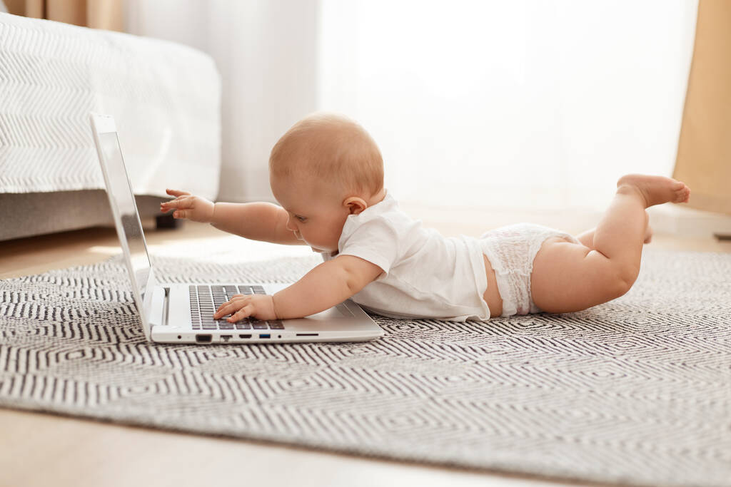Indoor shot cute ciekawa dziewczynka lub chłopiec w białej koszuli leżącej na podłodze na brzuchu, na szarym dywanie i dotykając laptopa, pozowanie samodzielnie w pokoju światła. - Zdjęcie, obraz