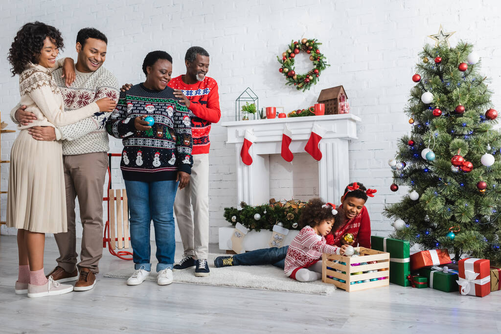 happy african american family looking at brother and sister playing near christmas tree in living room - Photo, Image