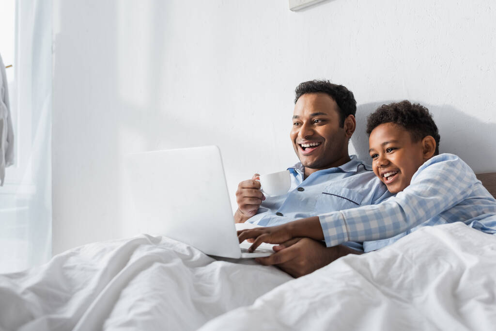 izgatott afro-amerikai fiú és apa néz vígjáték laptopon reggel az ágyban - Fotó, kép