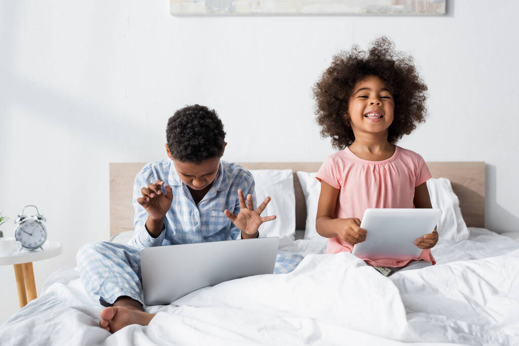 Afrikanisch-amerikanischer Junge mit Laptop im Bett neben fröhlicher Schwester mit digitalem Tablet - Foto, Bild