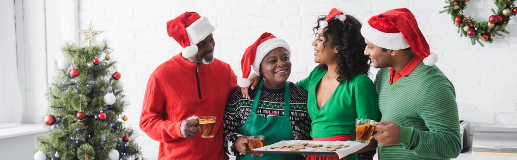 中年アフリカ系アメリカ人女性保持ベーキングシートとともにクリスマスクッキー近く幸せな家族とクリスマスツリー,バナー - 写真・画像