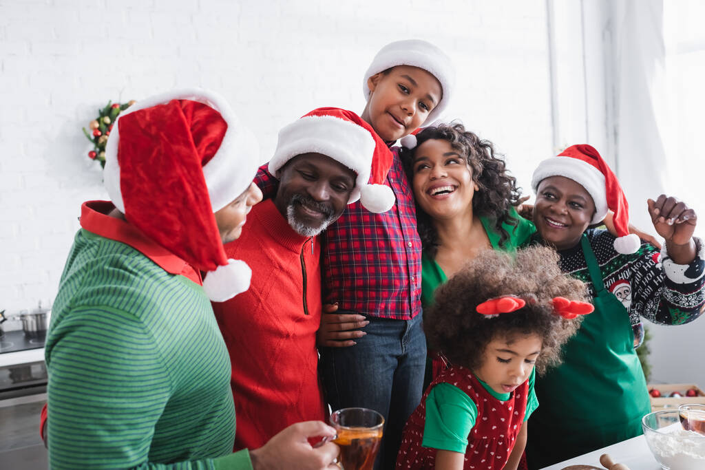 vidám afro-amerikai család télapó sapkában ölelkezve és mosolyogva a konyhában - Fotó, kép