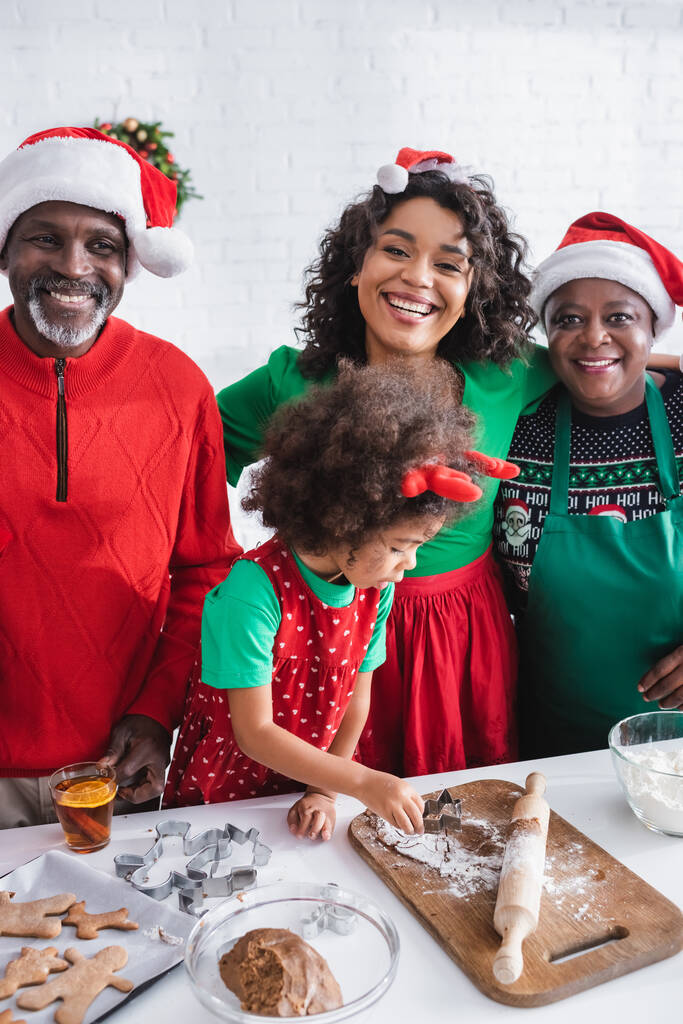 Αφροαμερικανή με κέρατα ταράνδου που ετοιμάζει χριστουγεννιάτικα μπισκότα κοντά στην ευτυχισμένη οικογένεια με καπέλα Σάντα - Φωτογραφία, εικόνα