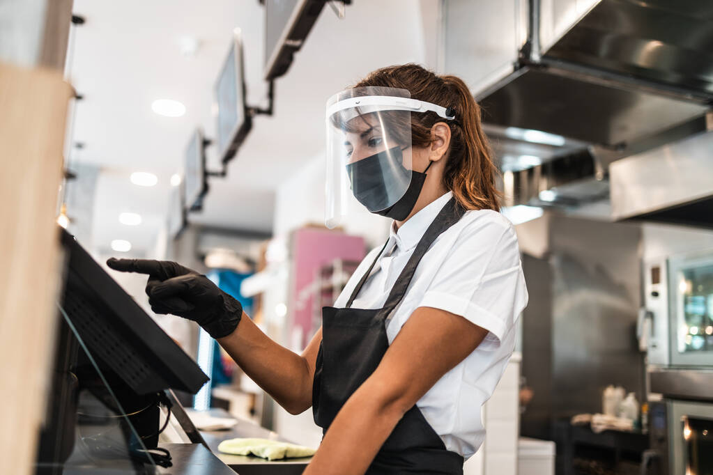 Beautiful young female worker with protective mask  and face shield working in bakery and using cache register machine. Business and coronavirus pandemic concept. - Photo, Image