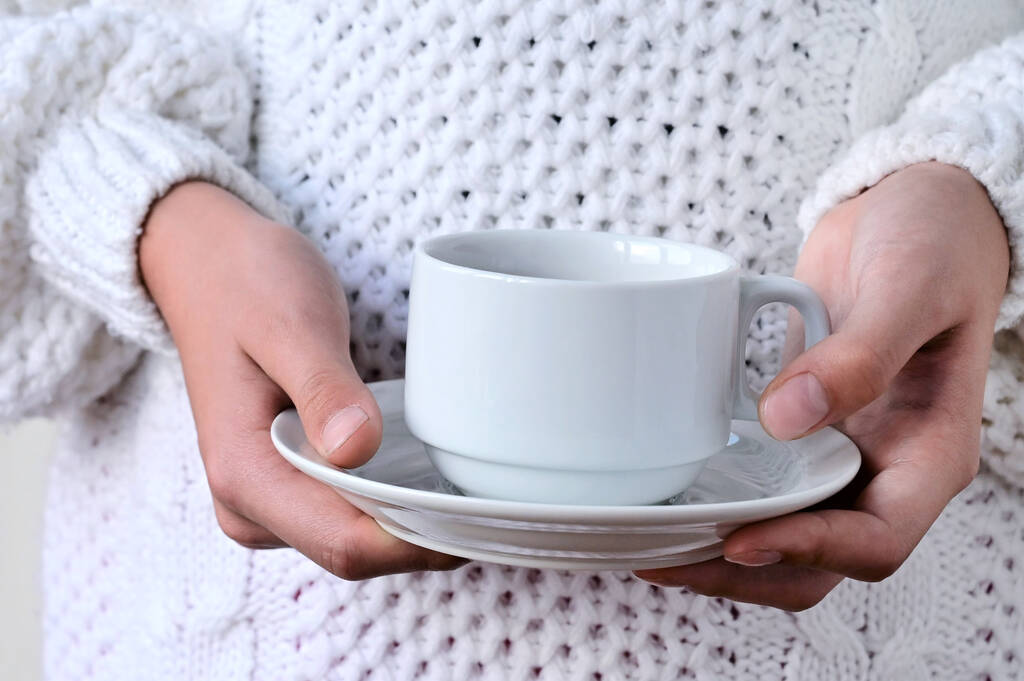 Beyaz fincan ve çay tabağı tutan, sıcak örülmüş beyaz kazaklı bir kızın ellerini yakın plan çek. Kış konforu, rahatlama kavramı - Fotoğraf, Görsel