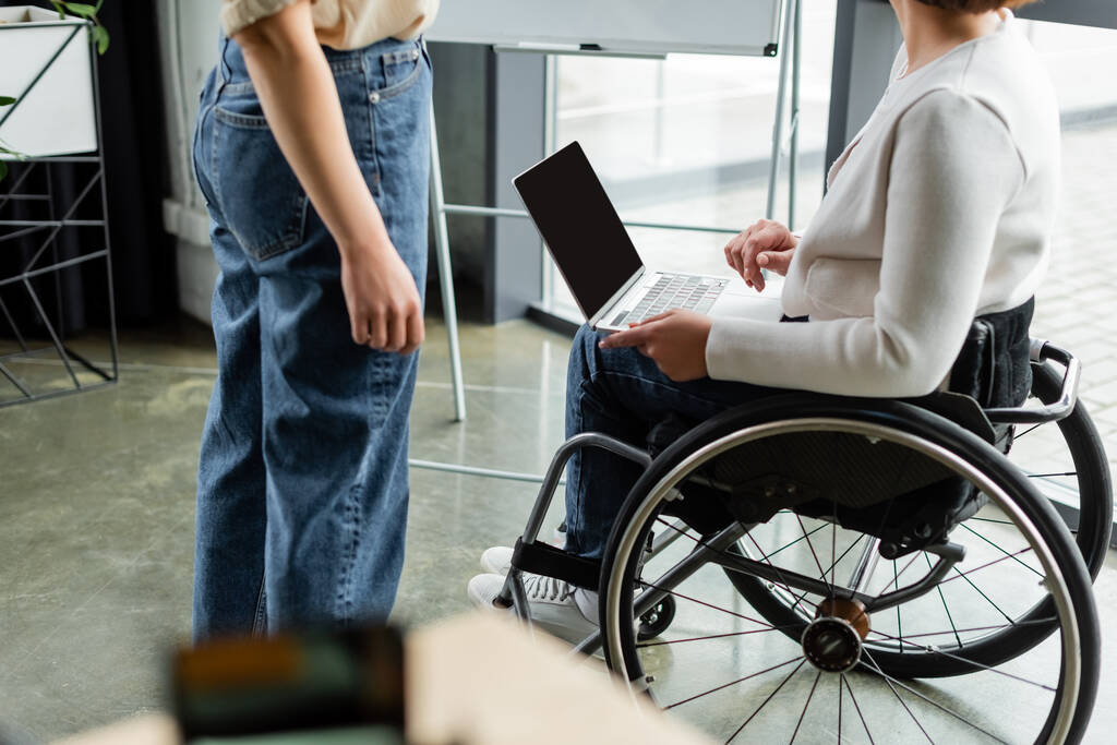 cropped view of businesswoman in wheelchair holding laptop with blank screen near colleague in office - Photo, Image