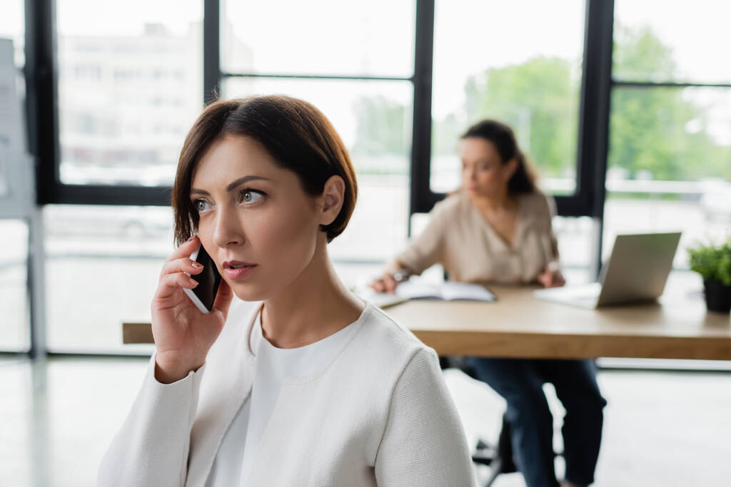 деловая женщина разговаривает по мобильному телефону в офисе рядом с африканским американским коллегой на размытом фоне - Фото, изображение