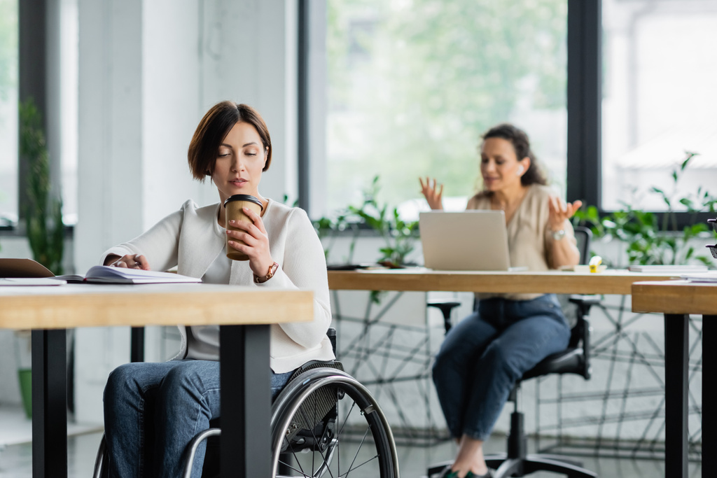businesswoman in wheelchair drinking coffee to go near african american colleague working on blurred background - Photo, Image