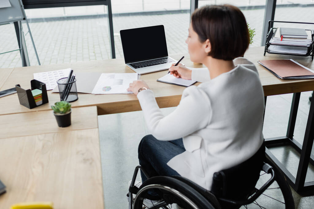 размытое деловая женщина в инвалидной коляске записи в ноутбуке во время работы с документами рядом с ноутбуком - Фото, изображение