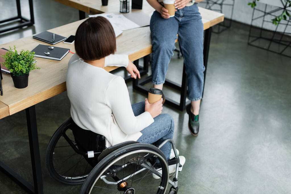 donna d'affari in sedia a rotelle in possesso di tazza di carta vicino collega seduto sulla scrivania durante la pausa caffè - Foto, immagini