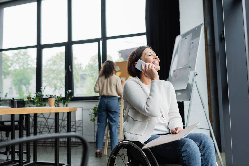 feliz mujer de negocios en silla de ruedas hablando en el teléfono inteligente cerca borrosa colega afroamericano en la oficina - Foto, Imagen
