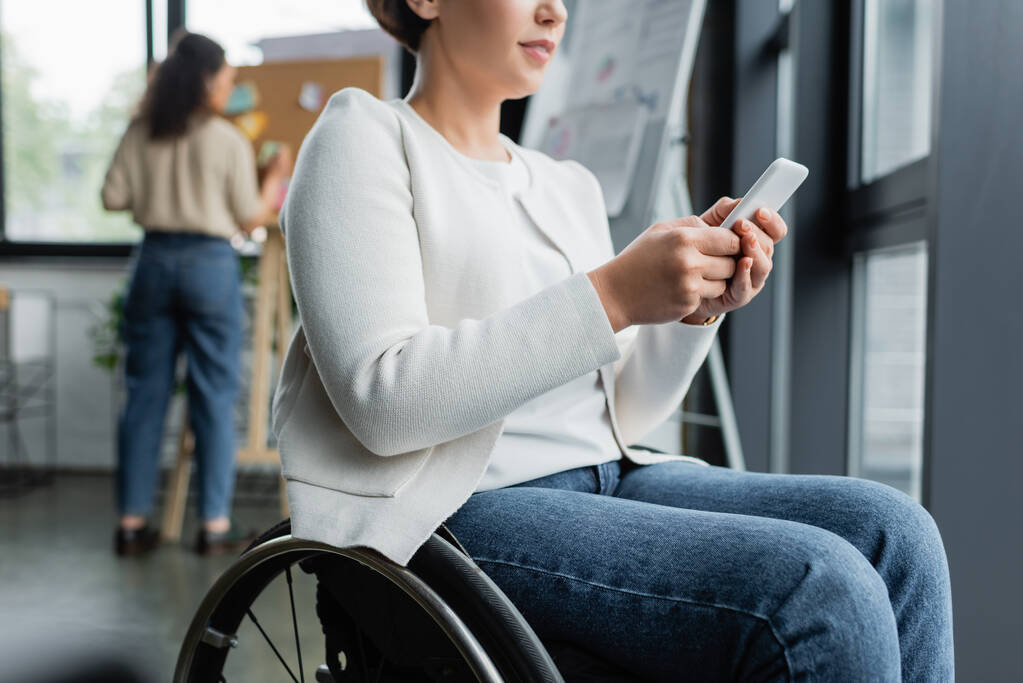 vista recortada de la mujer de negocios en la mensajería en silla de ruedas en el teléfono móvil cerca de un colega afroamericano en un fondo borroso - Foto, imagen