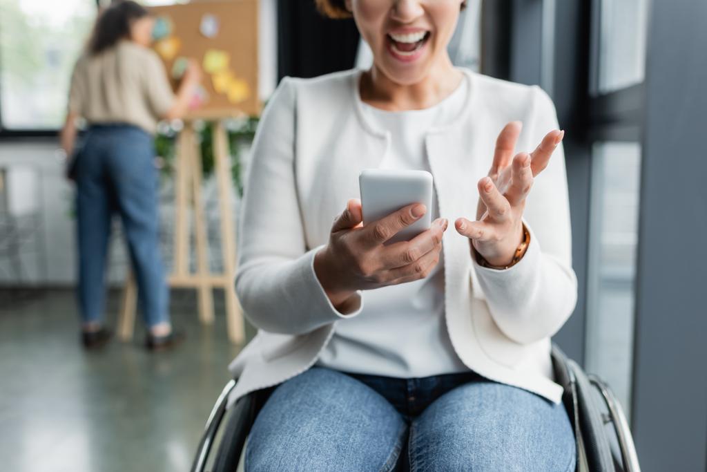 Wütende Geschäftsfrau im Rollstuhl benutzt Smartphone in der Nähe von afrikanisch-amerikanischem Kollegen, der an verschwommenem Hintergrund arbeitet - Foto, Bild