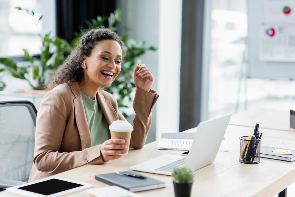 Αφροαμερικανή επιχειρηματίας με καφέ για να πάει χαμογελώντας κατά τη διάρκεια της βιντεοκλήσης στο φορητό υπολογιστή στο γραφείο - Φωτογραφία, εικόνα