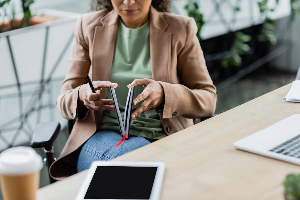 vista recortada de borrosa mujer de negocios afroamericana apertura portátil cerca de tableta digital en el lugar de trabajo - Foto, imagen