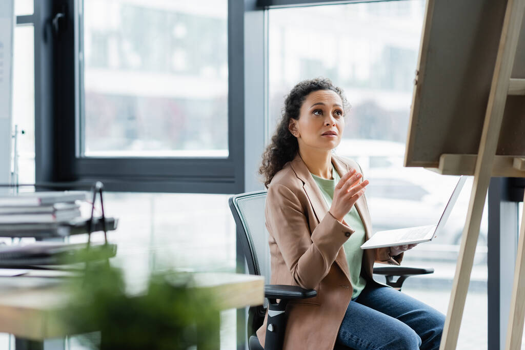 mulher de negócios afro-americana sentada com laptop e apontando para o quadro de notas no escritório - Foto, Imagem