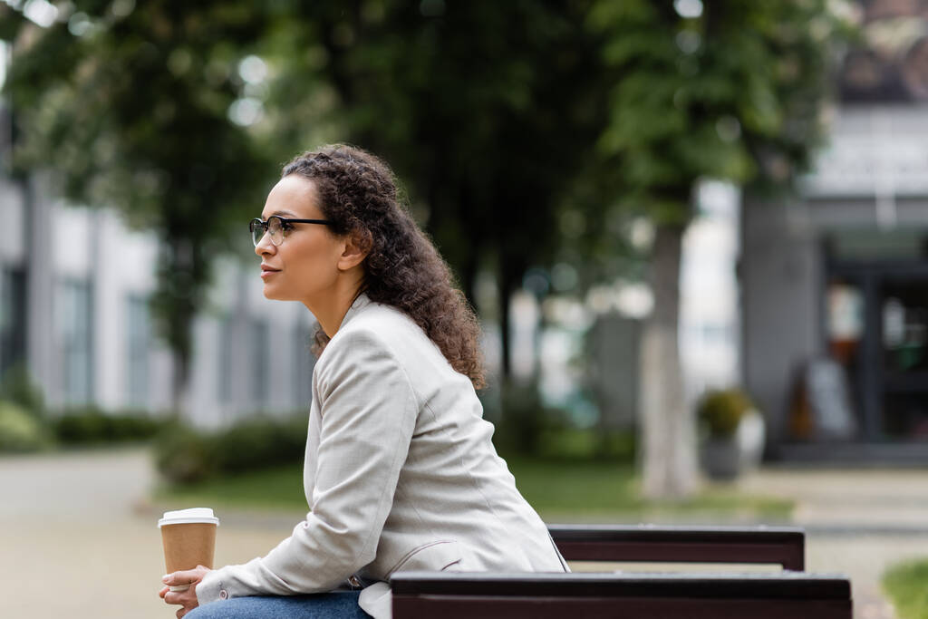 вид сбоку африканской бизнесвумен в очках, сидящей с кофе, чтобы выйти на улицу - Фото, изображение