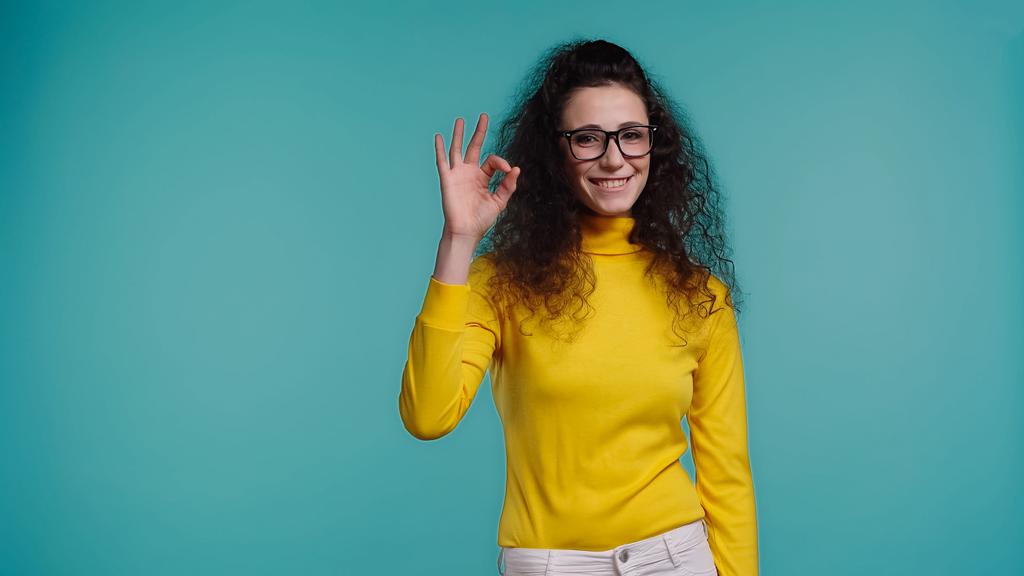 heureuse jeune femme dans des lunettes montrant ok signe isolé sur bleu  - Photo, image
