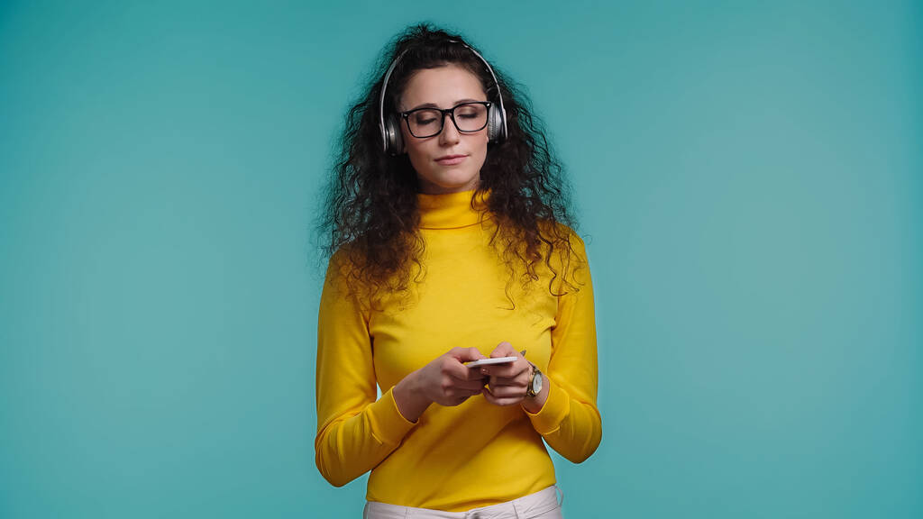 junge Frau mit Brille hält Smartphone in der Hand, während sie Musik im Kopfhörer hört, isoliert auf blauem Grund  - Foto, Bild