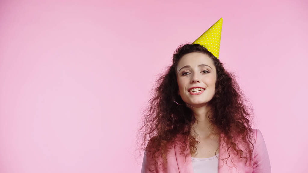 feliz joven rizado mujer en partido gorra felicitando con cumpleaños aislado en rosa  - Foto, Imagen