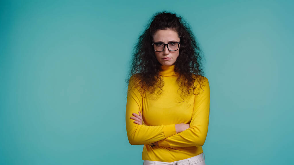 jeune femme frustrée dans des lunettes et col roulé debout avec les bras croisés isolés sur bleu - Photo, image
