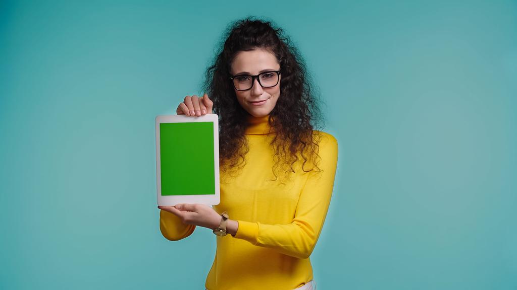 青で隔絶された緑の画面のデジタルタブレットを持つメガネの若い陽気な女性  - 写真・画像
